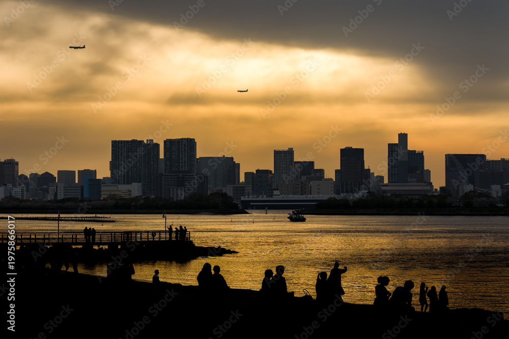 Naklejka premium Silhouetted people relaxing on Odaiba beach during a golden sunset over Tokyo Bay