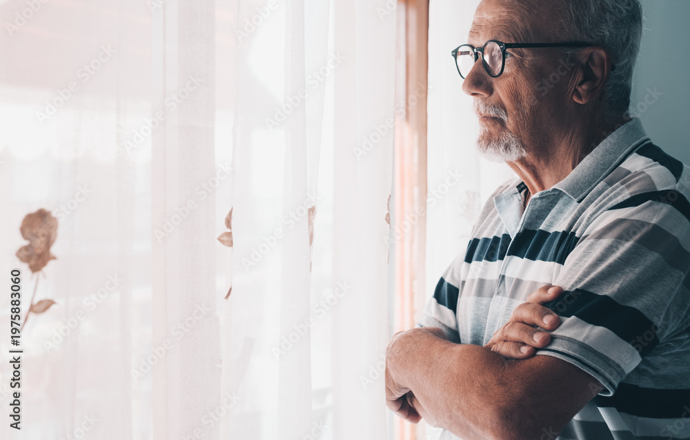 Fototapeta premium Older man in striped polo standing by window looking thoughtful and reflective at home