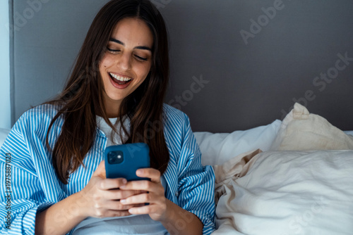 Smiling woman in striped shirt using a smartphone in bed, relaxing and messaging at home