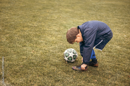 Tablou pe pânză A young boy football player tying the shoelace on his soccer cleat on a backyard