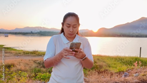 Asian woman using smartphone while relaxing at the lake during sunset with mountain background