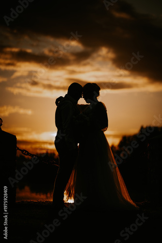 Silhouette of a couple embracing during sunset, with a dramatic sky and water reflection creating a romantic atmosphere in an outdoor setting