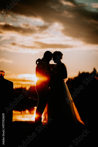 Silhouette of a couple embracing during sunset, with a vibrant sky and reflections on the water creating a romantic atmosphere in an outdoor setting