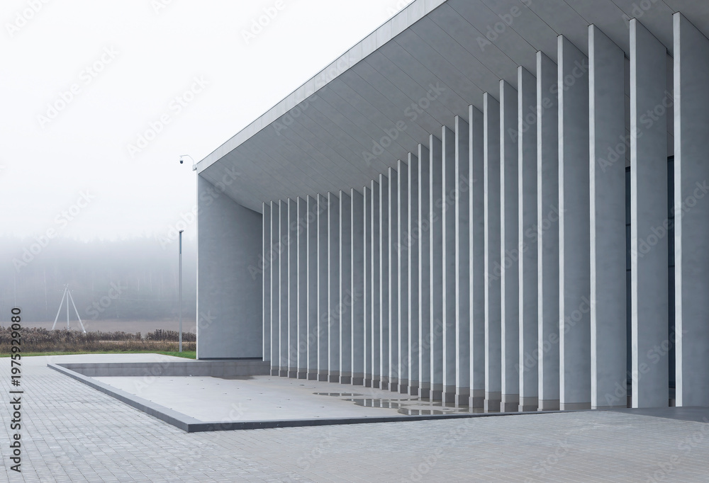 Fototapeta premium Curved colonnade of modern building stands beside empty paved space and open landscape. Repeating vertical columns create smooth architectural rhythm along exterior. Concept of public space, minimal