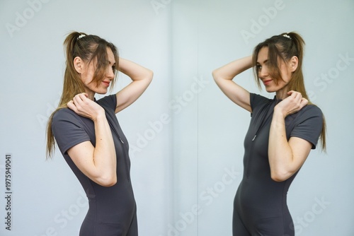 Young woman touching hair while looking at mirror reflection. Gray fitted bodysuit, minimal interior, soft light, calm lifestyle moment, symmetry composition.