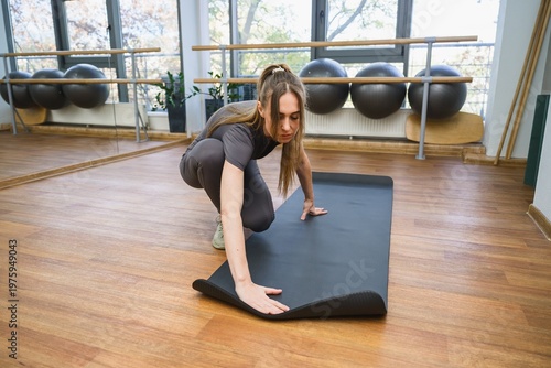 Young woman unrolling yoga mat in bright fitness studio. Preparing for workout, healthy lifestyle concept, gym interior with mirrors and equipment.
