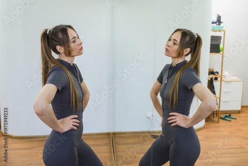 Young woman standing in fitness studio looking at her reflection in mirror. Body awareness, self evaluation and training progress concept.