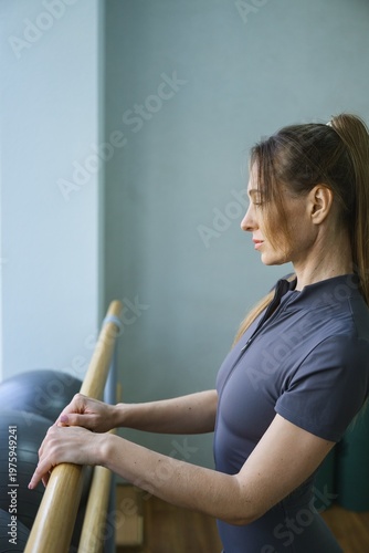 Young woman standing at ballet barre with eyes closed in fitness studio. Relaxation, mindfulness and body awareness during training, calm wellness concept.