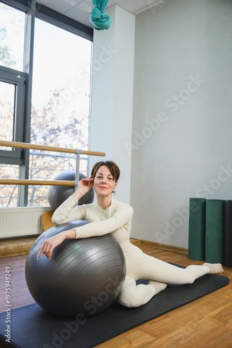 Young woman sitting on mat leaning on fitness ball and smiling at camera. Relaxation after training, wellness, healthy lifestyle and recovery concept.