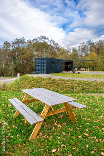 Wooden picnic table in front of modern lounge bar structure built from repurposed shipping containers, an outdoor terrace with seating arrangements and covered patio in peaceful natural green setting