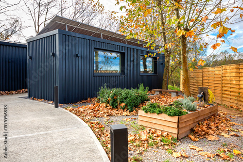 Modern eco friendly coworking office with dark facade and rooftop solar panels, pathway and raised garden beds with autumn leaves showcasing eco friendly building in nature