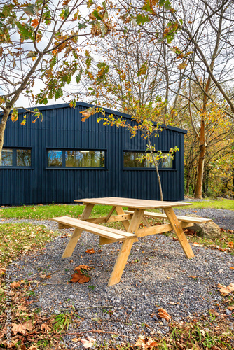 Modern and recycled dark gray corrugated metal cabin with large windows and a wooden picnic table on a gravel and grass area, surrounded by trees with autumn leaves in a peaceful natural park setting