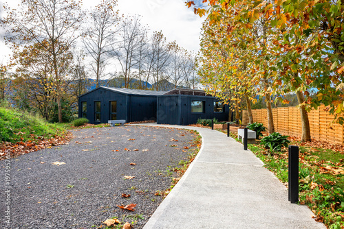 Modern home built with shipping containers featuring a dark exterior and solar panels on the roof, surrounded by a gravel driveway, concrete pathway, and autumn trees with colorful leaves