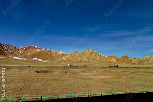 A truck traveling along a highway through a vast arid mountain landscape under a clear deep blue sky, evoking wide open space, transport, travel, solitude, and remote infrastructure concepts
