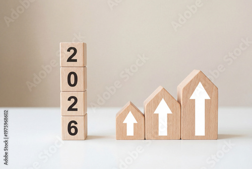 Wooden toy houses with white upward arrows next to wooden blocks spelling 2026 on white desk symbolizing real estate market growth property investment and rising housing prices