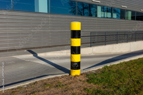 Fotografie A vertical yellow and black striped safety bollard stands prominently in front of a modern industrial building, marking the edge of a concrete ramp under bright daylight