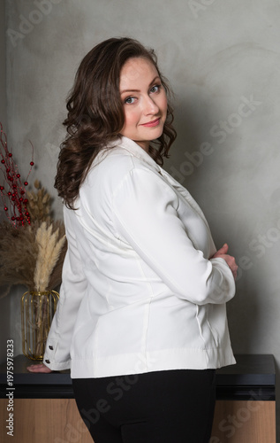 Back view of pretty brunette lady smiling at camera. Caucasian woman with long wavy brown hair wearing a white jacket looking back over her shoulder with a gentle smile while posing in modern office