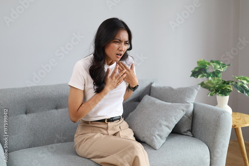 Fotografie A young woman sits on a sofa at home holding her chest, feeling intense pain, shortness of breath, panic attack, heart attack, stroke