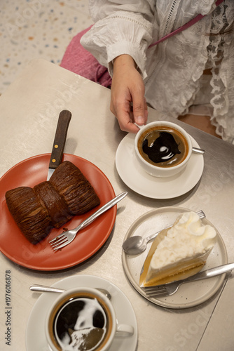 Aesthetic overhead view of a woman's hand holding a black coffee cup next to a chocolate bow-tie croissant and a slice of coconut cream cake on a modern metallic café table.