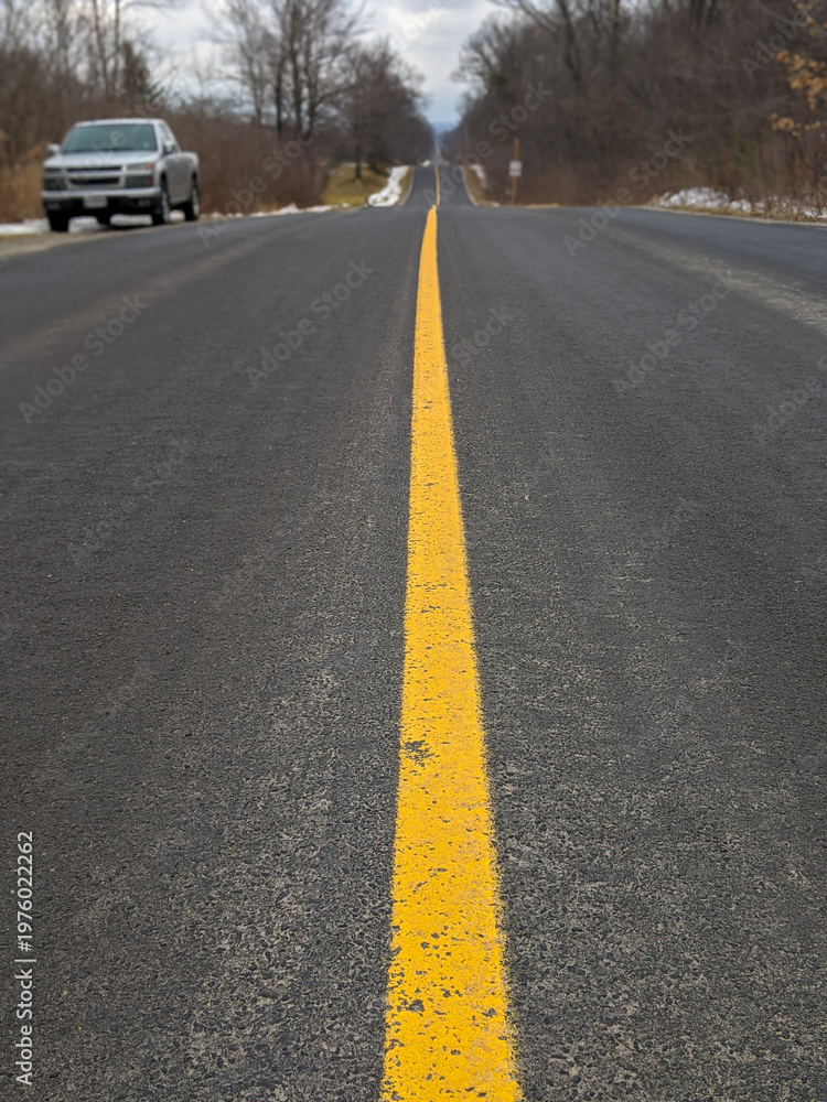 Obraz premium A pickup truck parked to the side of an empty rural road stretching to the horizon, dominated by a bright yellow center line. Bordered by bare winter trees beneath an overcast sky, evoking travel.