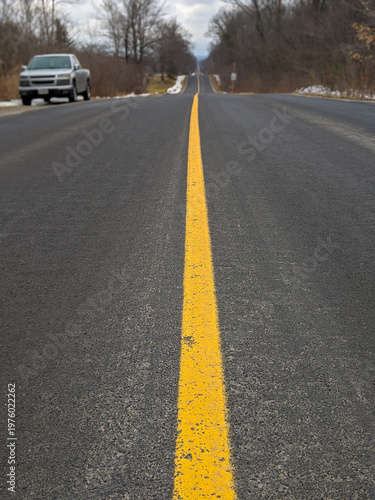 A pickup truck parked to the side of an empty rural road stretching to the horizon, dominated by a bright yellow center line. Bordered by bare winter trees beneath an overcast sky, evoking travel.