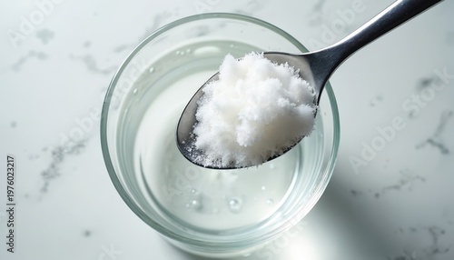 Spoon with white powder nears clear liquid in glass. Granules begin to dissolve, creating swirls. Preparation of drink or supplement ingredient.