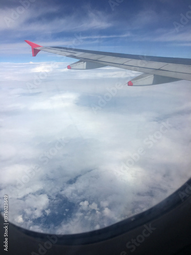 View of an airplane wing cutting across a sea of clouds, seen through a passenger window. Calm sky tones evoke travel, freedom, adventure and aerial perspective during a daytime flight.