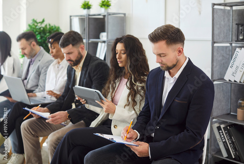 Employees, corporate staff members, applicants sitting in row in office working on diverse devices, writing notes, ideas in clipboard, preparing for job interview or upcoming meeting with clients
