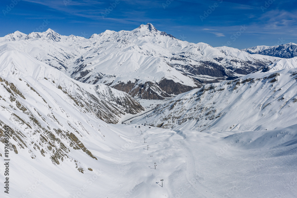 Fototapeta premium Aerial view of Mount Kazbek sunny winter day with snow covered mountains and clear sky, Kazbegi region near Gudauri, Georgia. Mount Kazbek bright sunlight and clear sky in Caucasus Mountains, Georgia