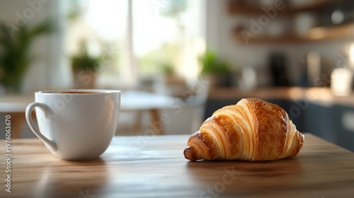 A croissant and a white cup of coffee sitting on a wooden table in a cosy kitchen provide an appetising backdrop for breakfast