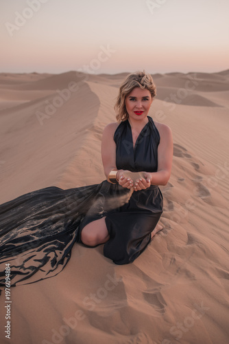 Beautiful woman sitting on sand dunes in elegant black dress.