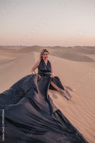 Beautiful woman sitting on sand dunes in elegant black dress.