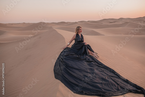 Beautiful woman sitting on sand dunes in elegant black dress.