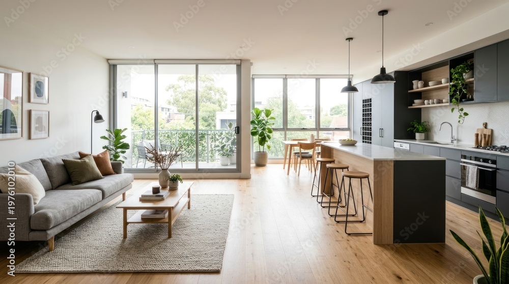 Fototapeta premium A bright and airy open-plan living area features a modern kitchen island and a comfortable living space. A gray sofa sits in front of a large window overlooking a city view.