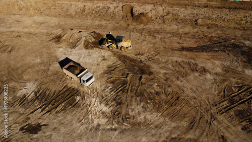 A high-angle aerial shot captures heavy machinery operating on a dirt construction site. A yellow front-end loader is shown filling the bed of a large dump truck with soil and debris.
