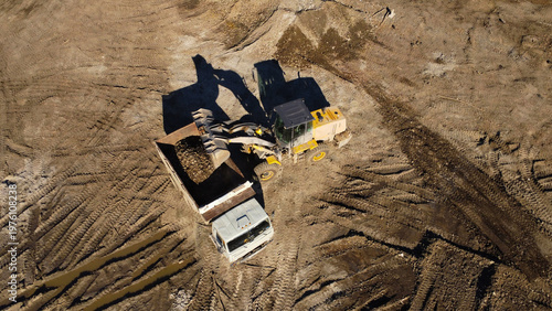 A high-angle aerial shot captures heavy machinery operating on a dirt construction site. A yellow front-end loader is shown filling the bed of a large dump truck with soil and debris.