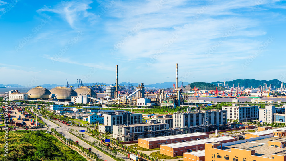 Obraz premium Modern industrial zone with large storage silos and factory buildings near the harbor under blue sky.