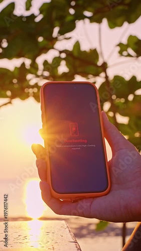 Vertical video. Smartphone Screen Showing Overheating Warning Message in Hand at Sunset with Tropical Background, Device Temperature Concept