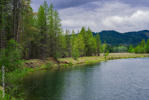 2021-06-14 THE COEUR D'ALENES RIVER WITH EVERGREENS LINING THE BANK WITH MOUNTAINS AND A CLOUDY SKY OUTSIDE OF HARRISON IDAHO