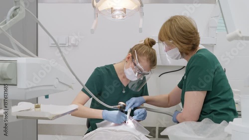 Dentist performing deep teeth cleaning with air flow sandblasting technology and assistant using saliva ejector. Medical team working in modern dental office for oral health and hygiene