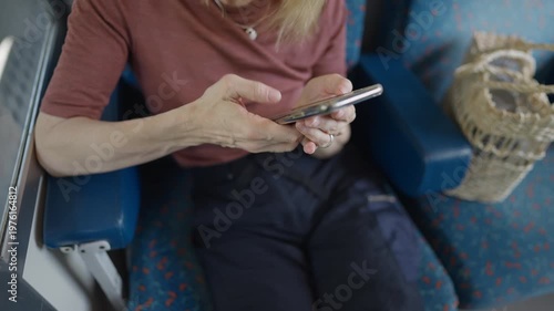 Mature blonde woman sitting by the window in a train, using her smartphone. She is typing and scrolling on her mobile device