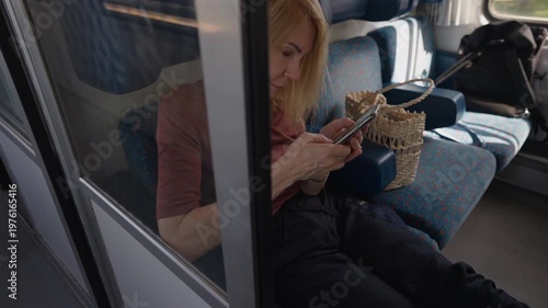 Mature woman sitting on a train seat by the window, engrossed in using her smartphone. Sunlight streams into the moving railway car