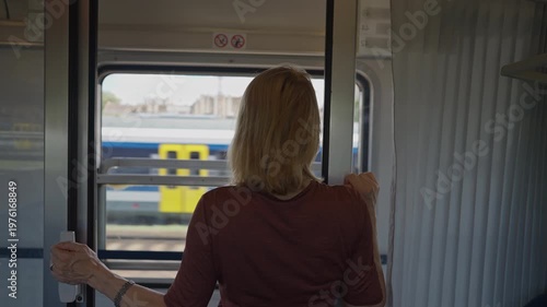 Rear view of a blonde woman standing in a train compartment, looking out the window at the passing landscape and other moving trains
