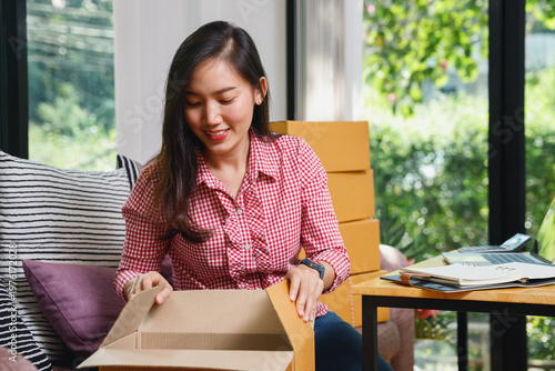 Happy young Asian woman small business owner smiling while packing a cardboard box at home. Female e-commerce seller preparing customer orders for shipping. Startup and online business concept.