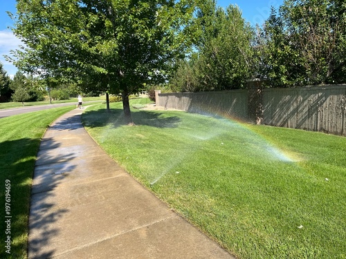 Φωτογραφία Lawn sprinklers spraying water across a green grassy area beside a sidewalk, creating a small rainbow in bright sunshine