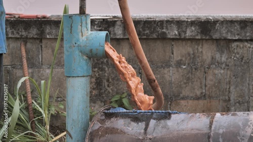 Worker manually drilling a groundwater well with muddy water flowing from a pipe outdoors. Suitable for construction, water supply, and rural development themes.