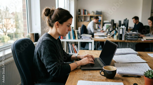 A woman with brown hair tied in a bun works on a laptop at a wooden desk in a modern office.