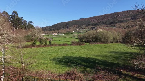 Green fields extending into a small village nestled below a forest-covered mountain under a blue sky