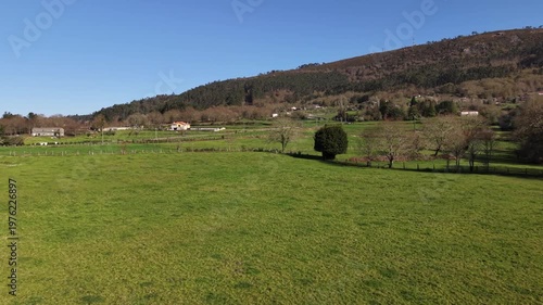 Green fields extending into a small village nestled below a forest-covered mountain under a blue sky