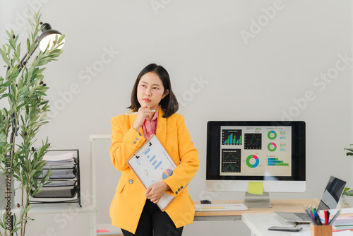 Introspective Moment: An office setting with an Asian businessperson contemplating strategy, holding documents, standing next to a computer displaying data analysis.